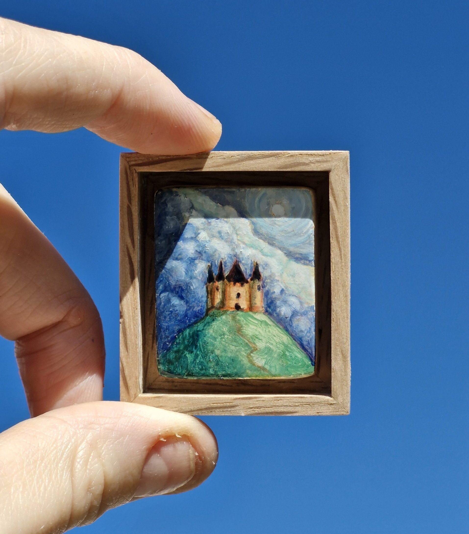peinture encadrée représentant un petit château. Elle est tenue dans une main. Le ciel bleu sans aucun nuage fait office de fond